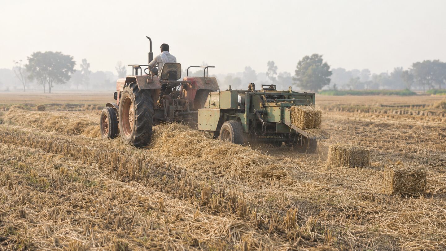 A tractor pulling a square baler through a harvested paddy field, with freshly formed straw bales dropping behind it